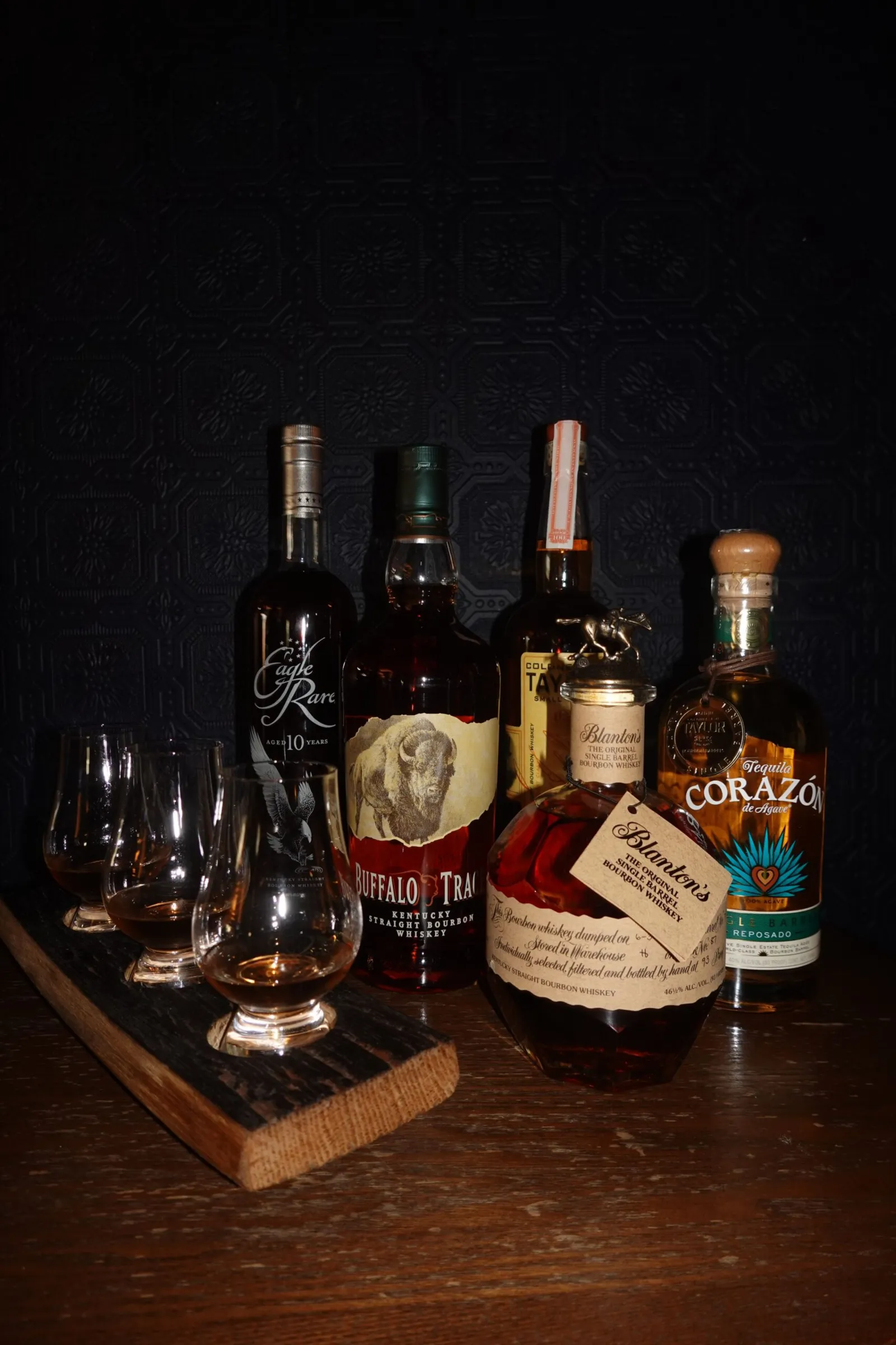 Five bottles of whiskey and tequila are arranged on a wooden surface, with four filled tasting glasses on a wooden tray in front—perfect for memorable events—set against a dark textured background.