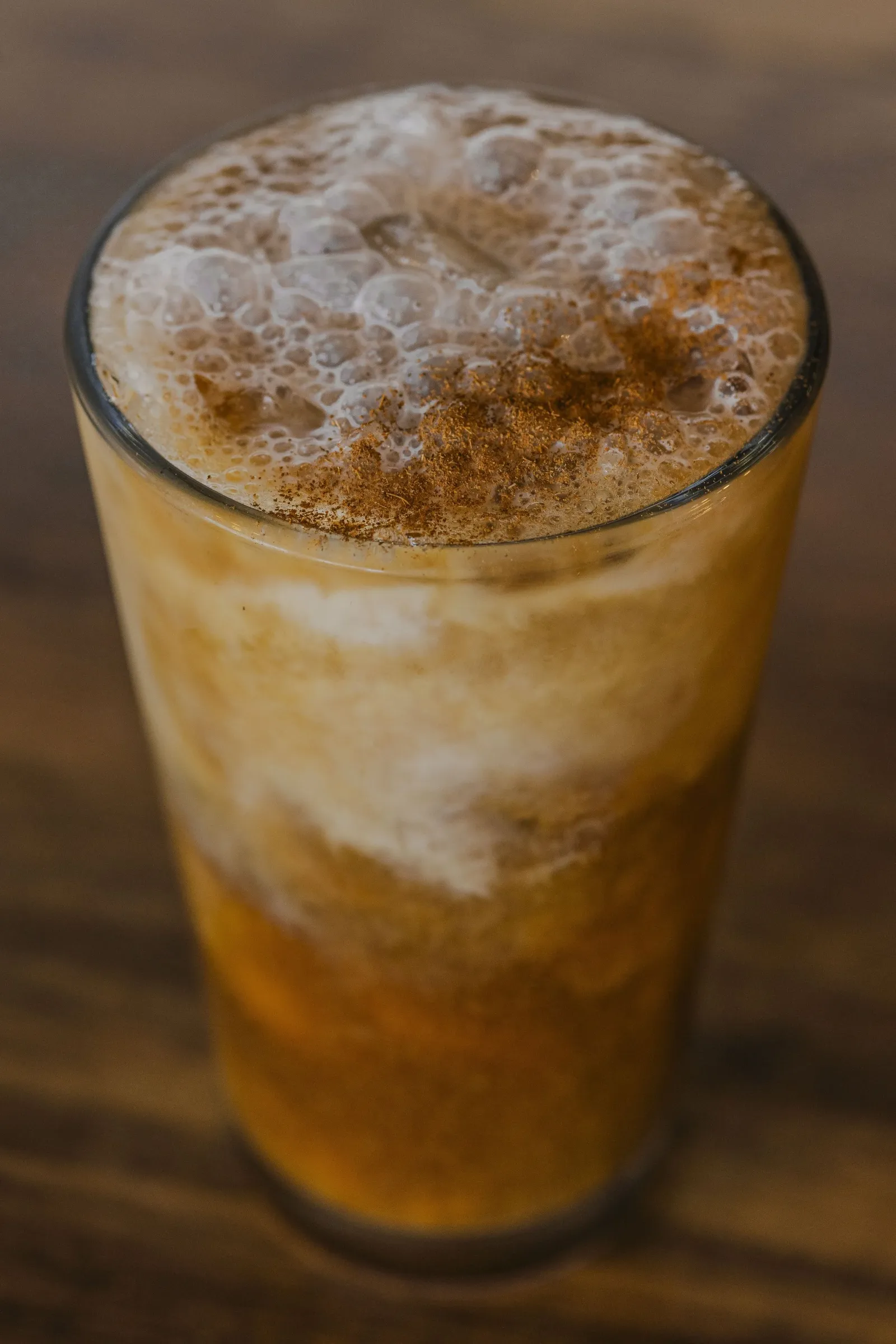 Iced kava coffee in a pint glass with white milk foam on top and a caramel colored liquid in the cup sitting on a wood table