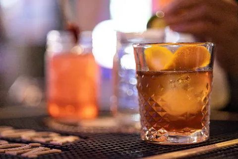 A glass of brown drink with orange slices, on a bar counter with blurred drinks in the background.