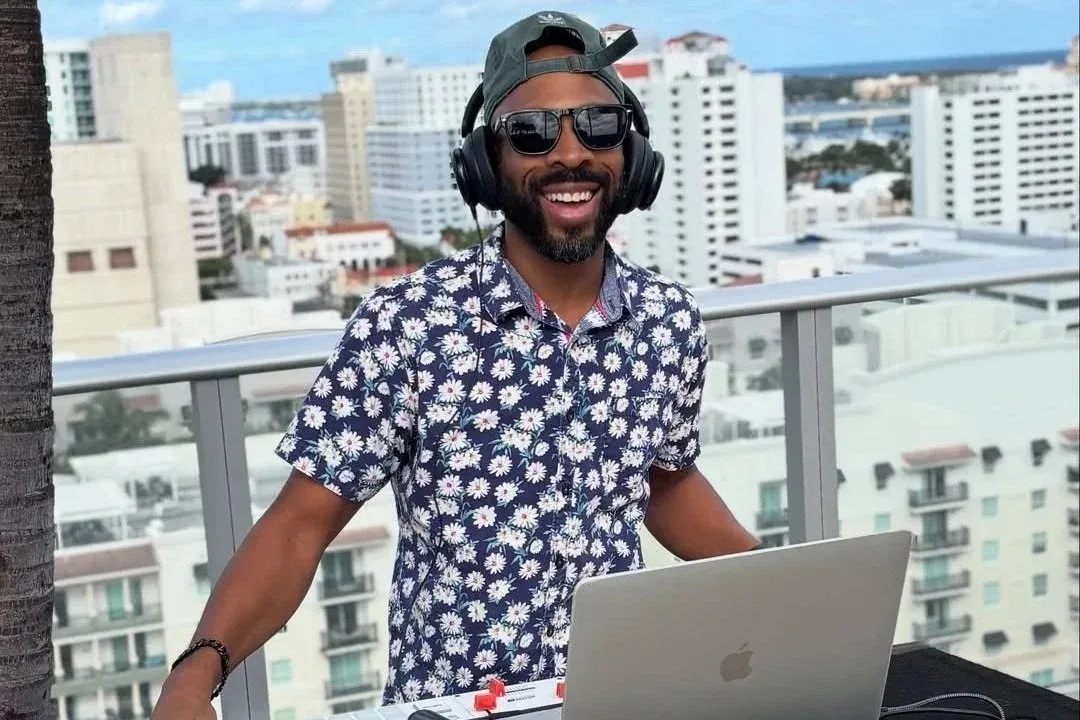 A man wearing sunglasses, a cap backwards, and large headphones, standing outdoors on a balcony with a cityscape background, smiling near a laptop.