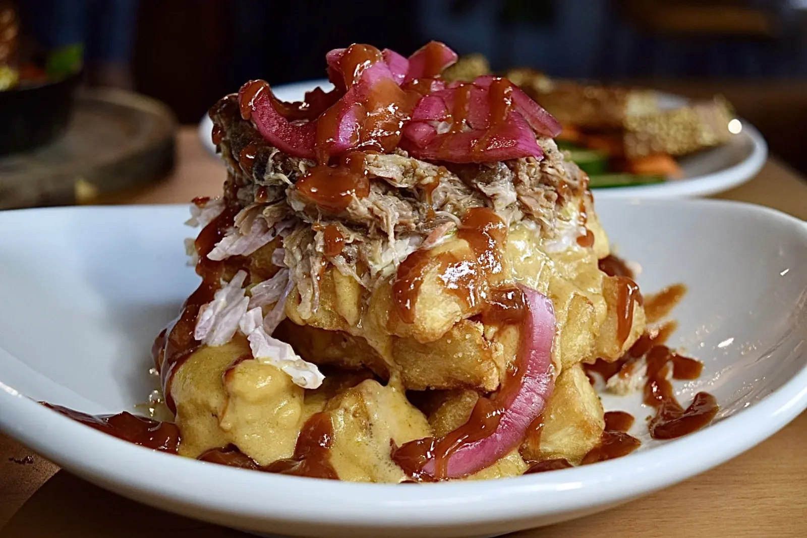A serving of loaded potato fries topped with shredded meat, chopped pickled red onions, and drizzled with barbecue sauce, in a white bowl.