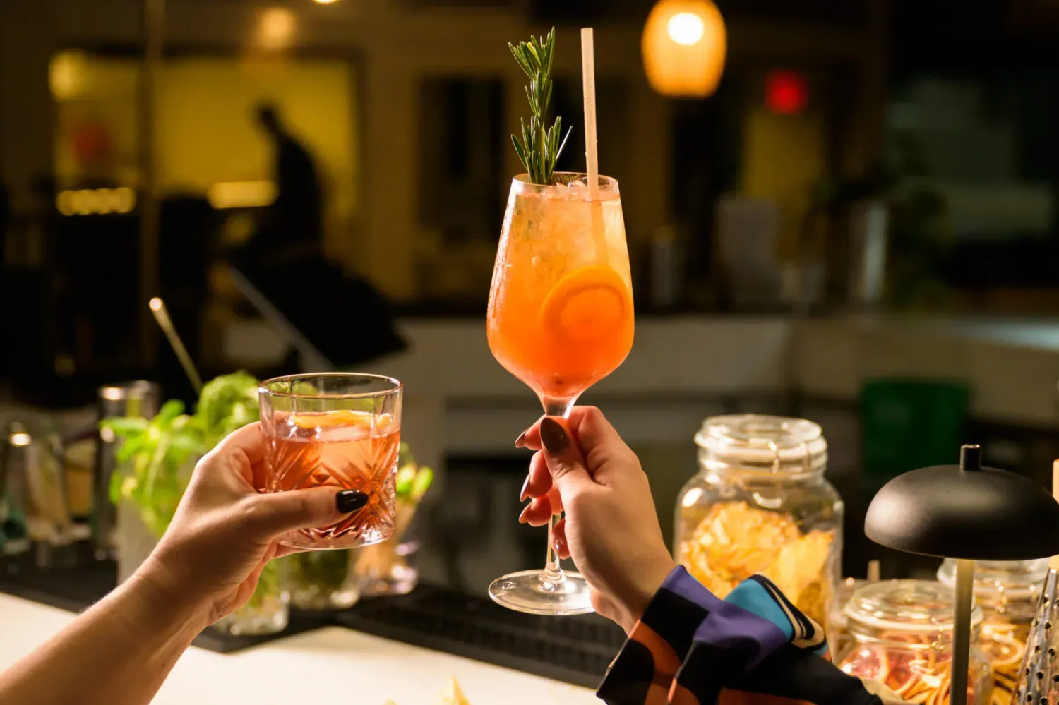 Two hands raising cocktails in a toast at an evening bar