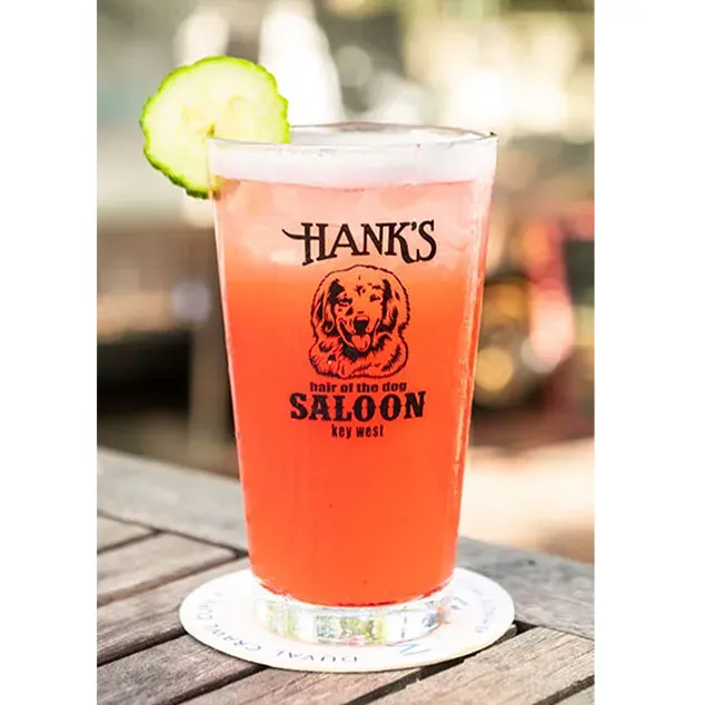 A pink cocktail in a Hank’s Saloon glass, garnished with a cucumber slice, sits on a wooden table—capturing the laid-back charm of a Key West bar.