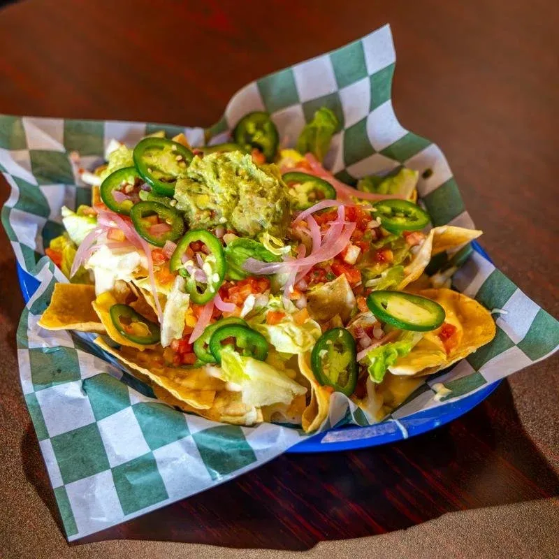 Nachos topped with jalapenos, guacamole, shredded lettuce, diced tomatoes, shredded cheese, sour cream, and sliced onions in a basket lined with checkered paper.