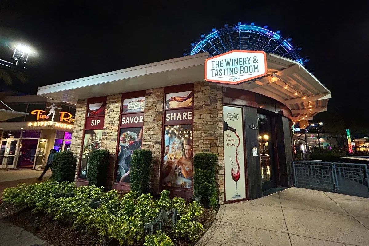 Night view of The Winery & Tasting Room with large neon sign, stone and glass exterior, surrounded by greenery, and a large Ferris wheel lit up in the background.