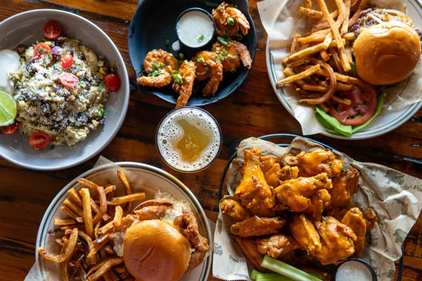 Assorted dishes of food including chicken wings with dipping sauce, French fries with a burger, a salad with cherry tomatoes, and fried chicken in a basket, all placed on a wooden table.