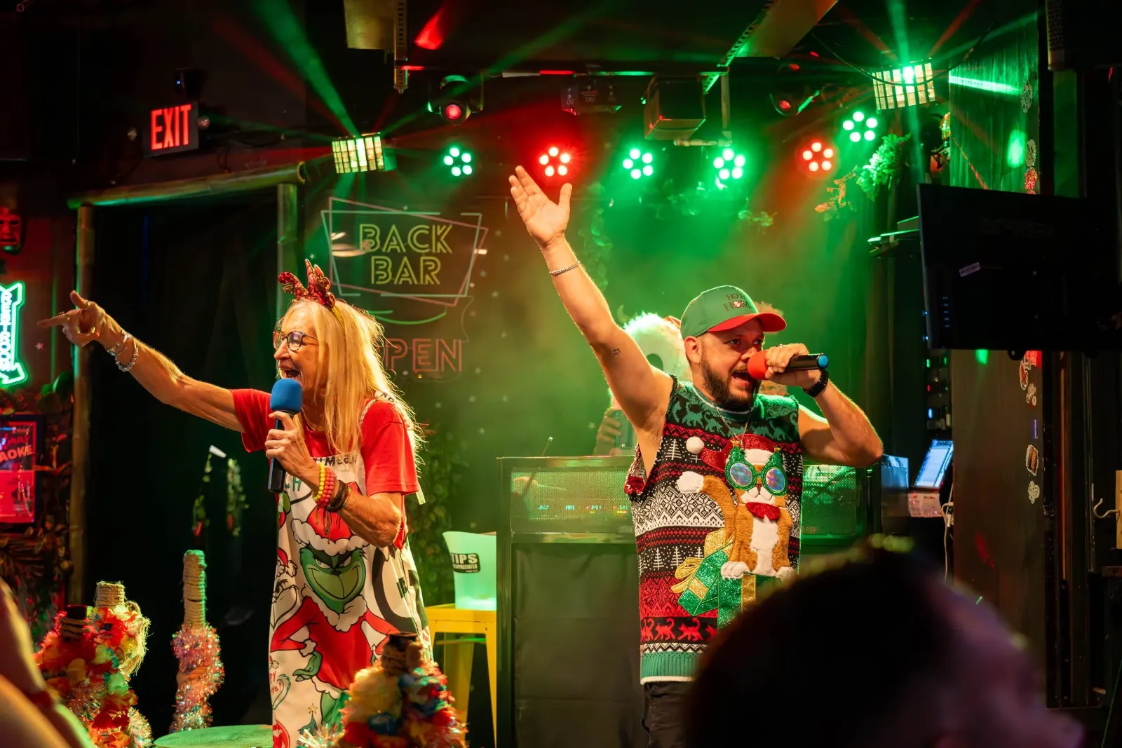 A woman and a man singing karaoke in a colorful, festive bar decorated with Christmas items. The woman has long blonde hair, glasses, and is wearing a red shirt and holiday-themed dress with a headban