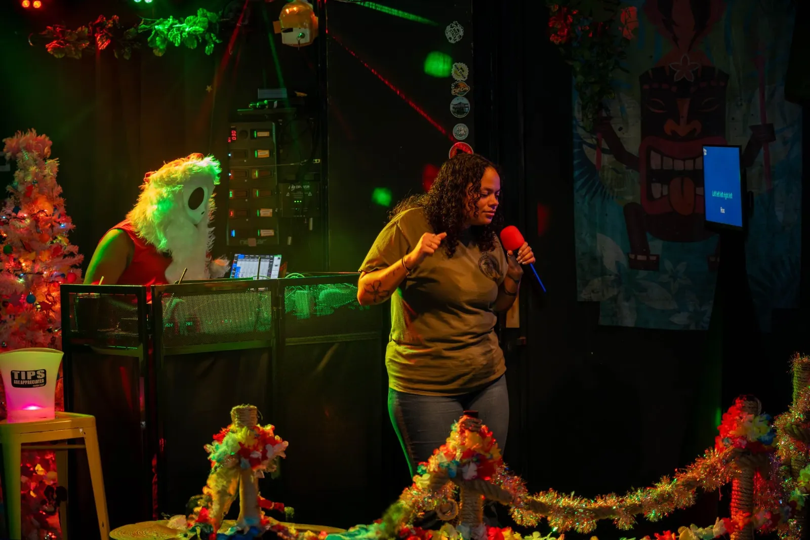 A woman singing into a red microphone in a decorated room with Christmas-themed decorations, including a Christmas tree and colorful garlands, while a DJ with a mask stands behind a DJ setup surrounde