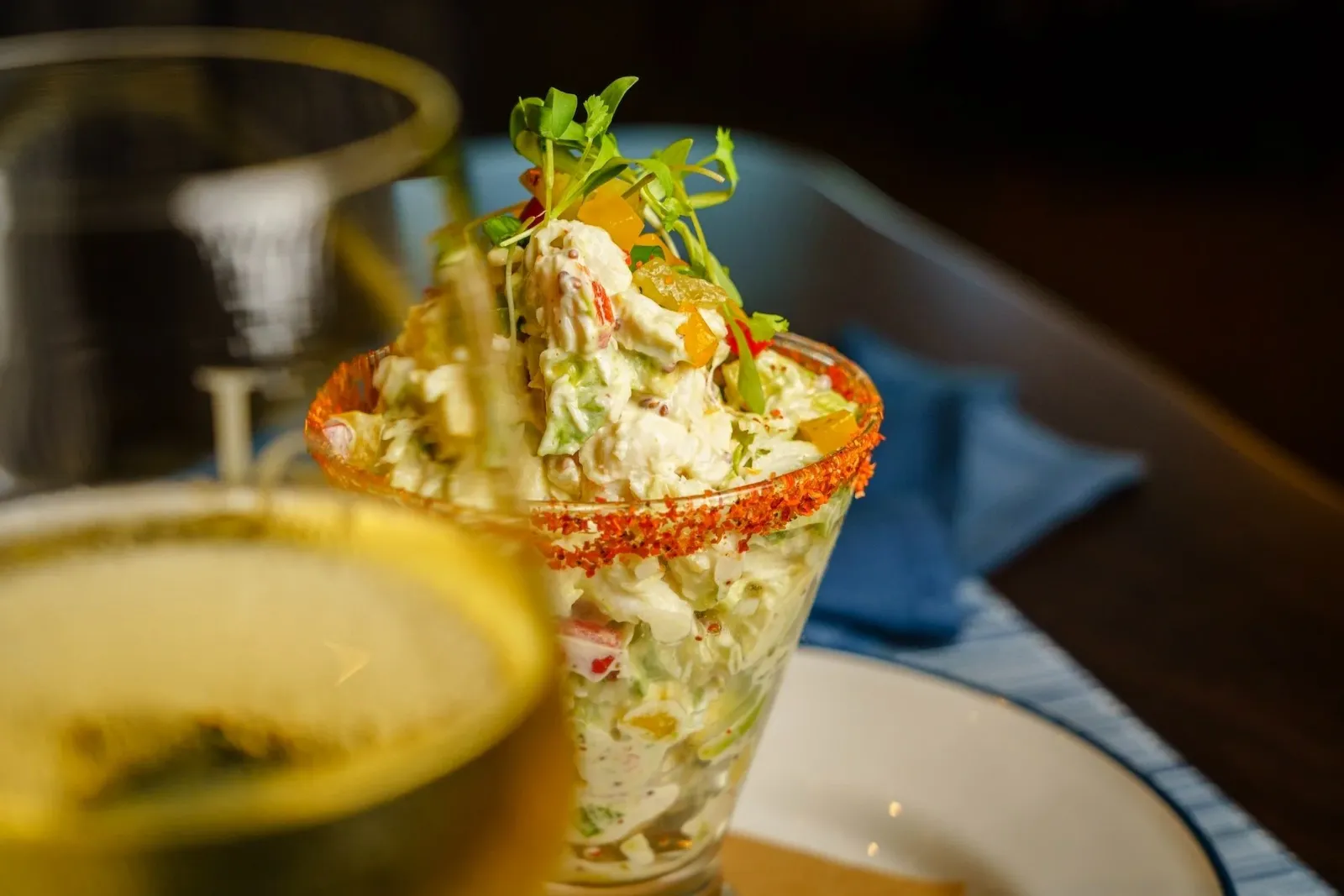 Close-up of a gourmet seafood salad served in a glass with a spiced rim, topped with fresh herbs and diced yellow peppers.