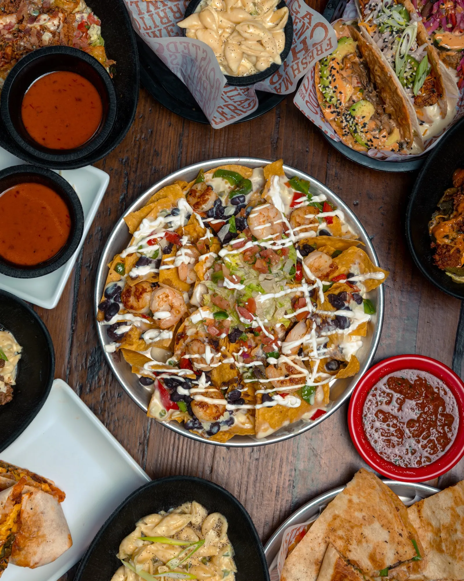 Assorted Mexican dishes on a wooden table including nachos topped with shrimp, black beans, sour cream, and vegetables, along with tacos, queso, and salsa.