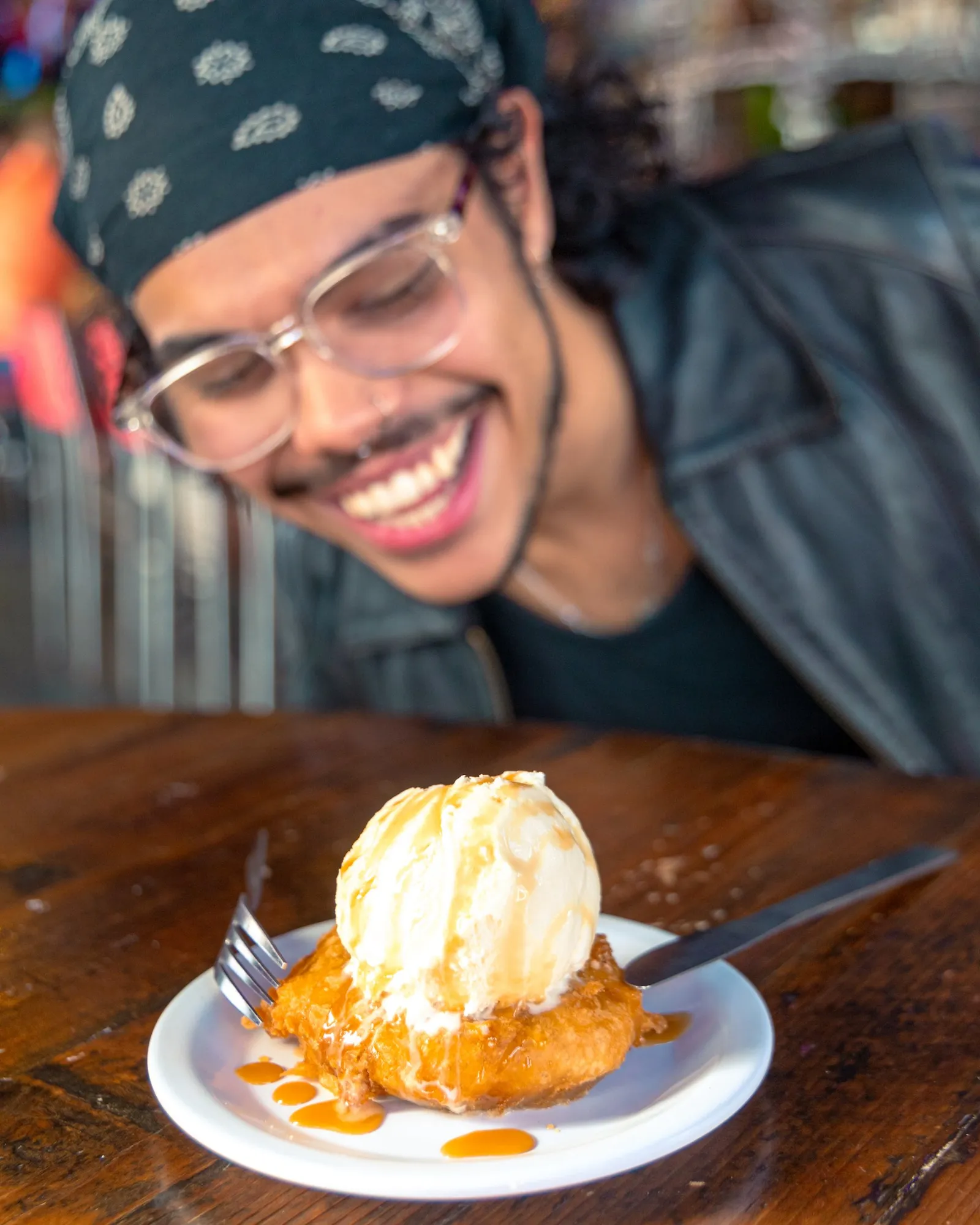 A person with glasses, a black bandana, and a leather jacket smiling at a plate of fried dessert topped with vanilla ice cream and caramel sauce.