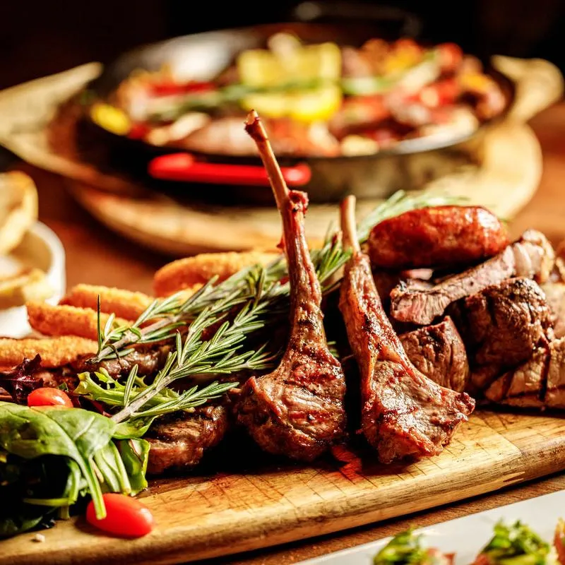 Grilled lamb chops, sausage, and steak on a wooden board, garnished with rosemary.