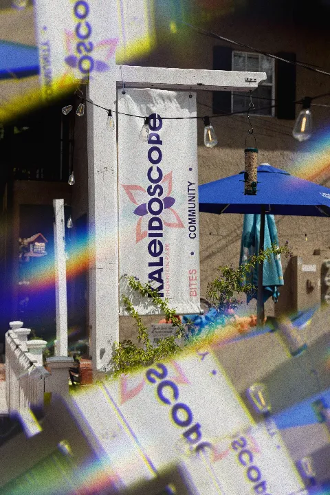 A sign for Kaleidoscope in Thornton Park in Orlando, Florida with purple and orange design, hanging on a white post outside a building, next to a blue patio umbrella, with string lights and bird feede