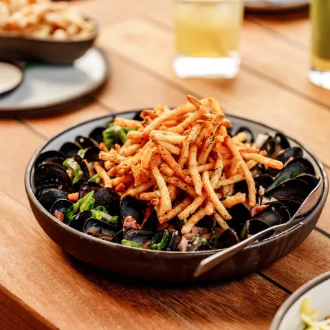 A plate of Moules Frites sits on a wooden table with other dishes and drinks.