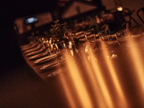 A long dining table set with plates, utensils, glasses, and a small plant centerpiece, illuminated by warm lighting.
