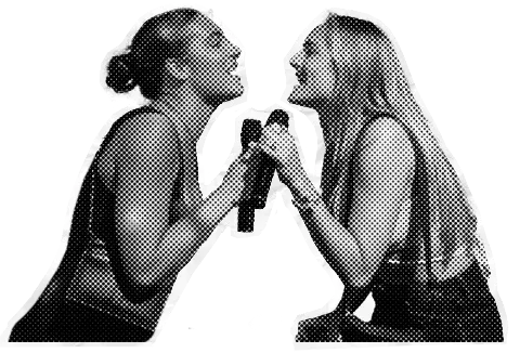 Two young women laughing and smiling at each other, holding bottles, with one making a gesture with her finger, in a playful and friendly interaction.