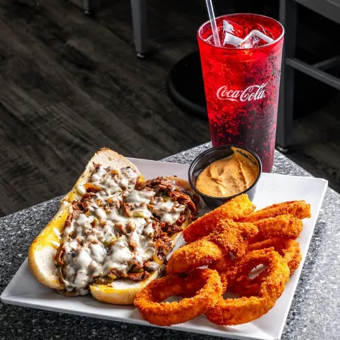 Philly Cheesesteak sandwich served with onion rings, dipping sauce and a glass of soda.
