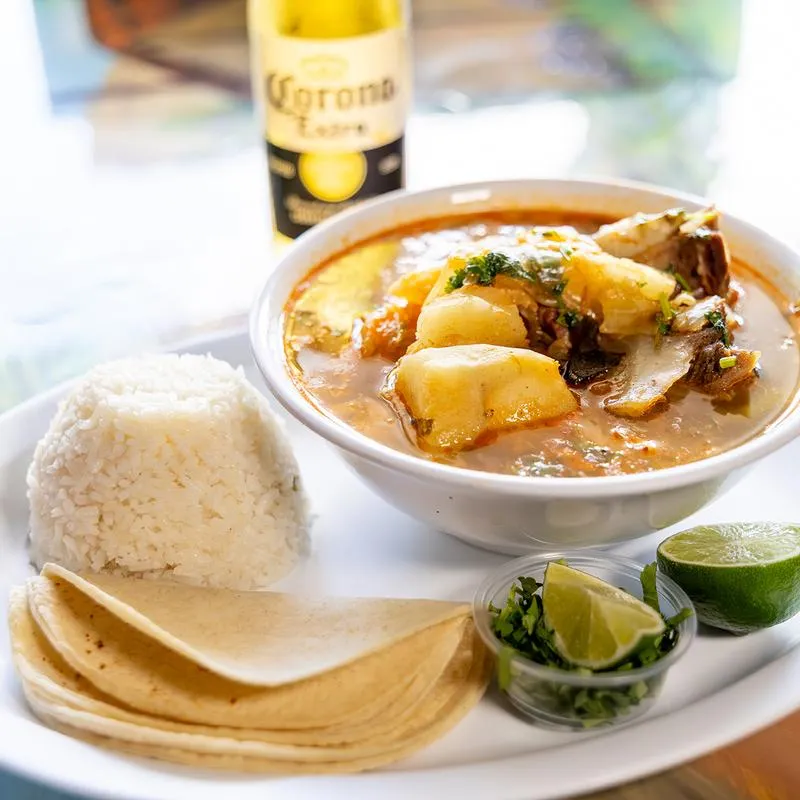 A bowl of seafood soup served with white rice, tortillas, and lime wedges.