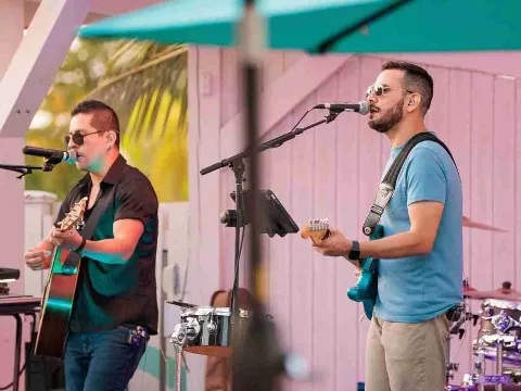Two men performing music outdoors; one playing guitar and singing into a microphone, the other playing guitar into a microphone; both wearing sunglasses.