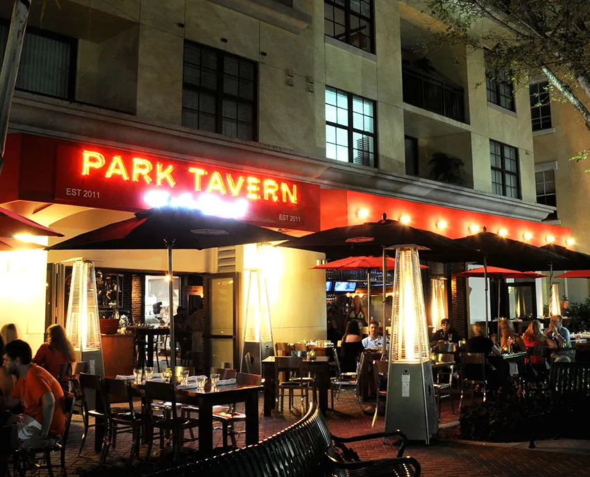 Park Tavern restaurant neon sign reading 'Est. 2011' above an outdoor patio with red umbrellas, patio heaters, and diners seated at night