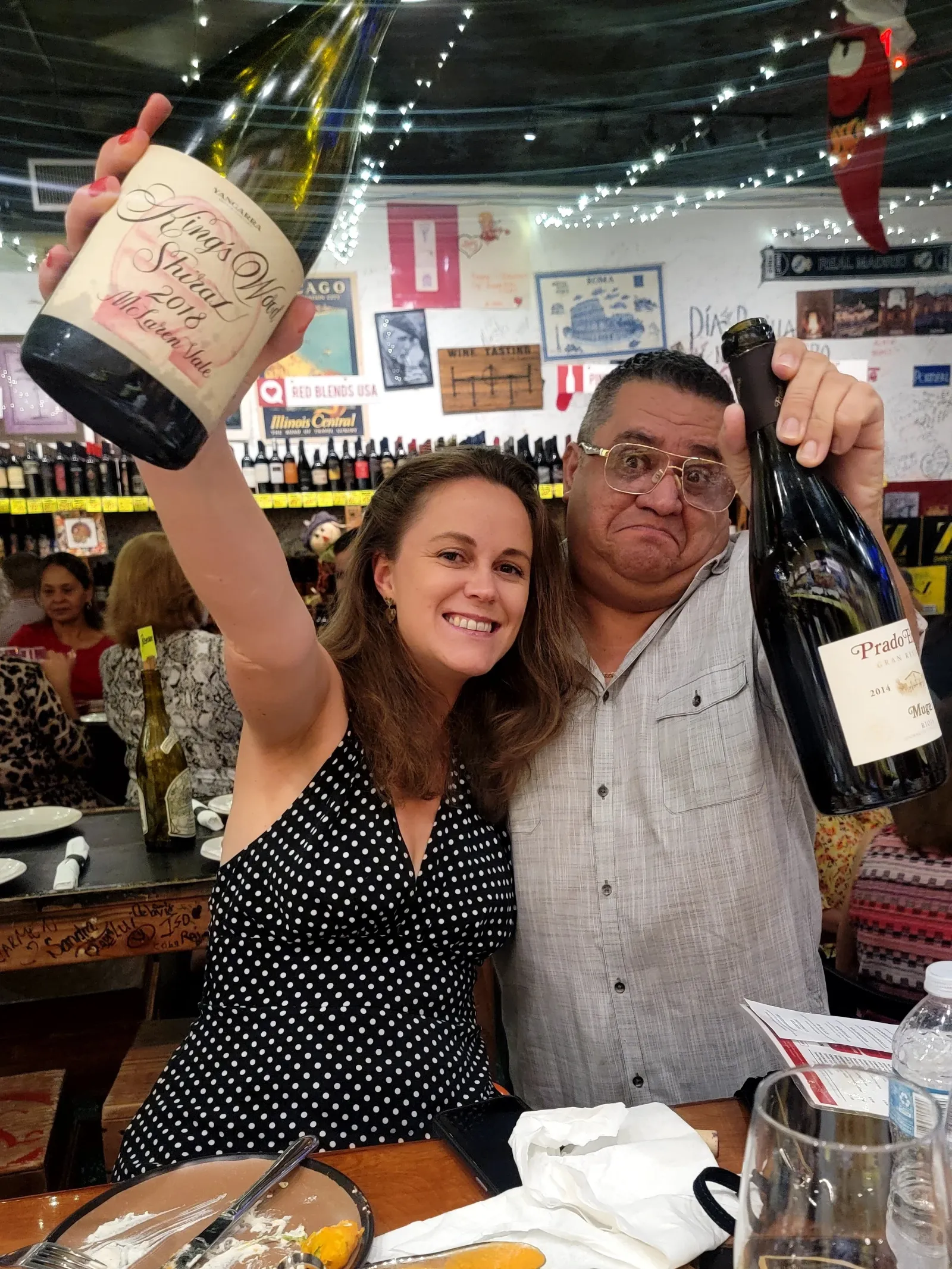 Two people celebrating at a restaurant, one woman holding a wine bottle with a custom label, both making faces for the camera. There are plates, utensils, and drinks on the table, with a decorated int