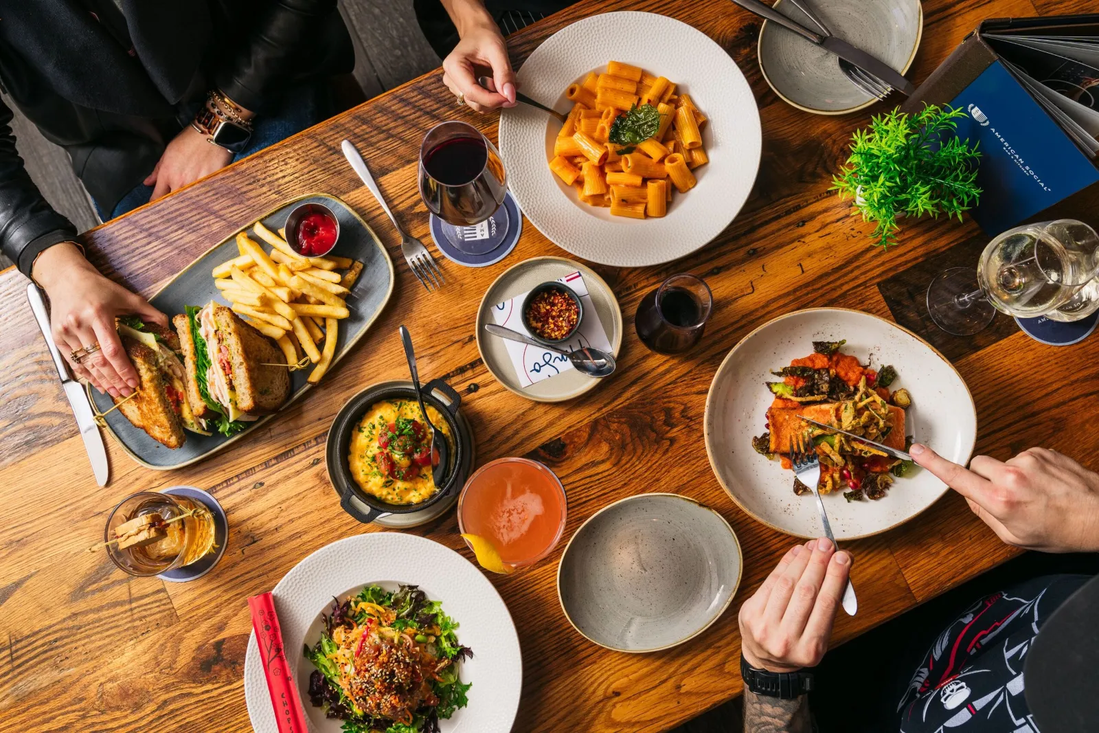 Overhead view of a wooden table set with plates of pasta, salad, grilled sandwiches with fries, a salmon dish, drinks, and small dishes from the menu, with three people reaching for food.