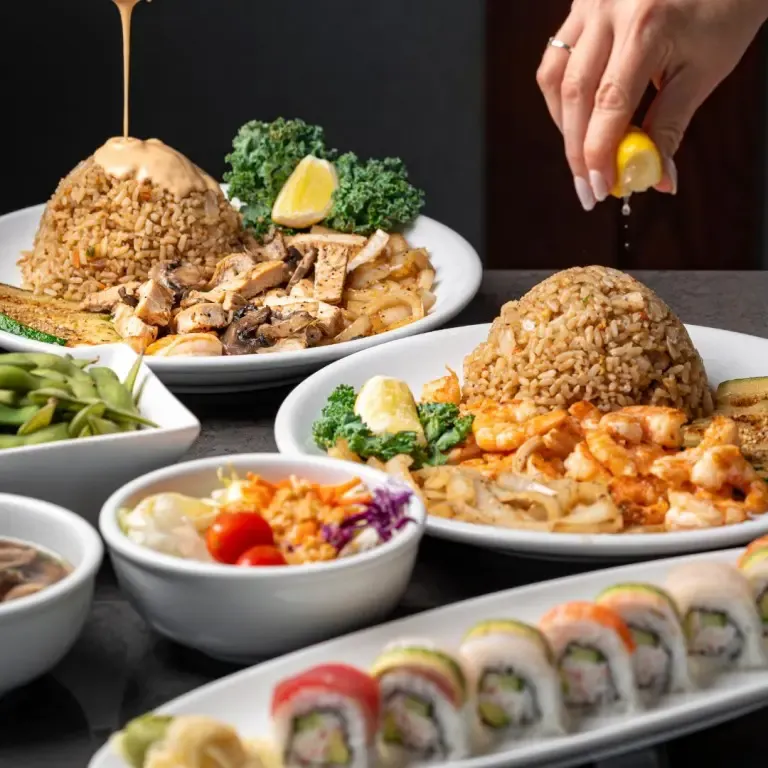 An assortment of Benihana dishes with hibachi rice, shrimp, chicken, sushi rolls, and edamame; hand squeezing lemon over rice.