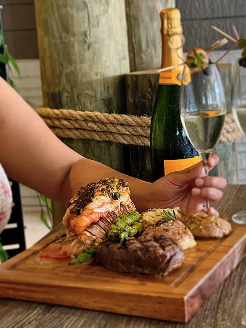Surf and turf platter with prime rib, a shrimp skewer, and potatoes, champagne bottle and glass in the background, hand holding the glass.