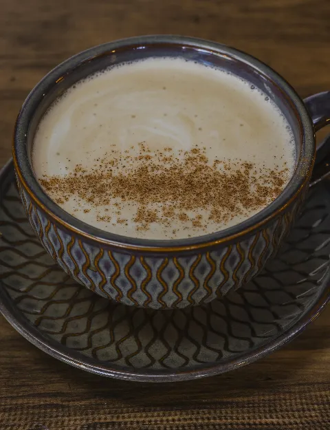 A cup of hot tea with milk and cinnamon floating on top, in a cup with a saucer sitting on a wood table