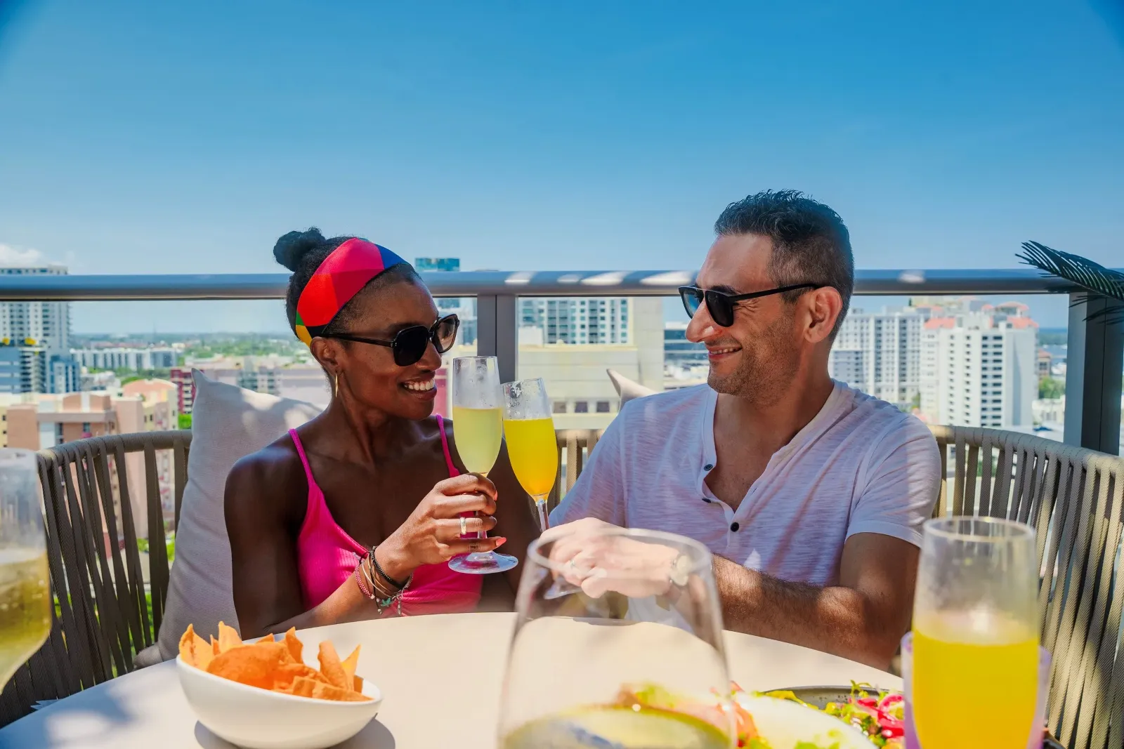 A man and woman on a rooftop terrace, holding champagne glasses, smiling, with a city skyline in the background. The woman has dark hair in a bun, wearing sunglasses and a pink top. The man has short 