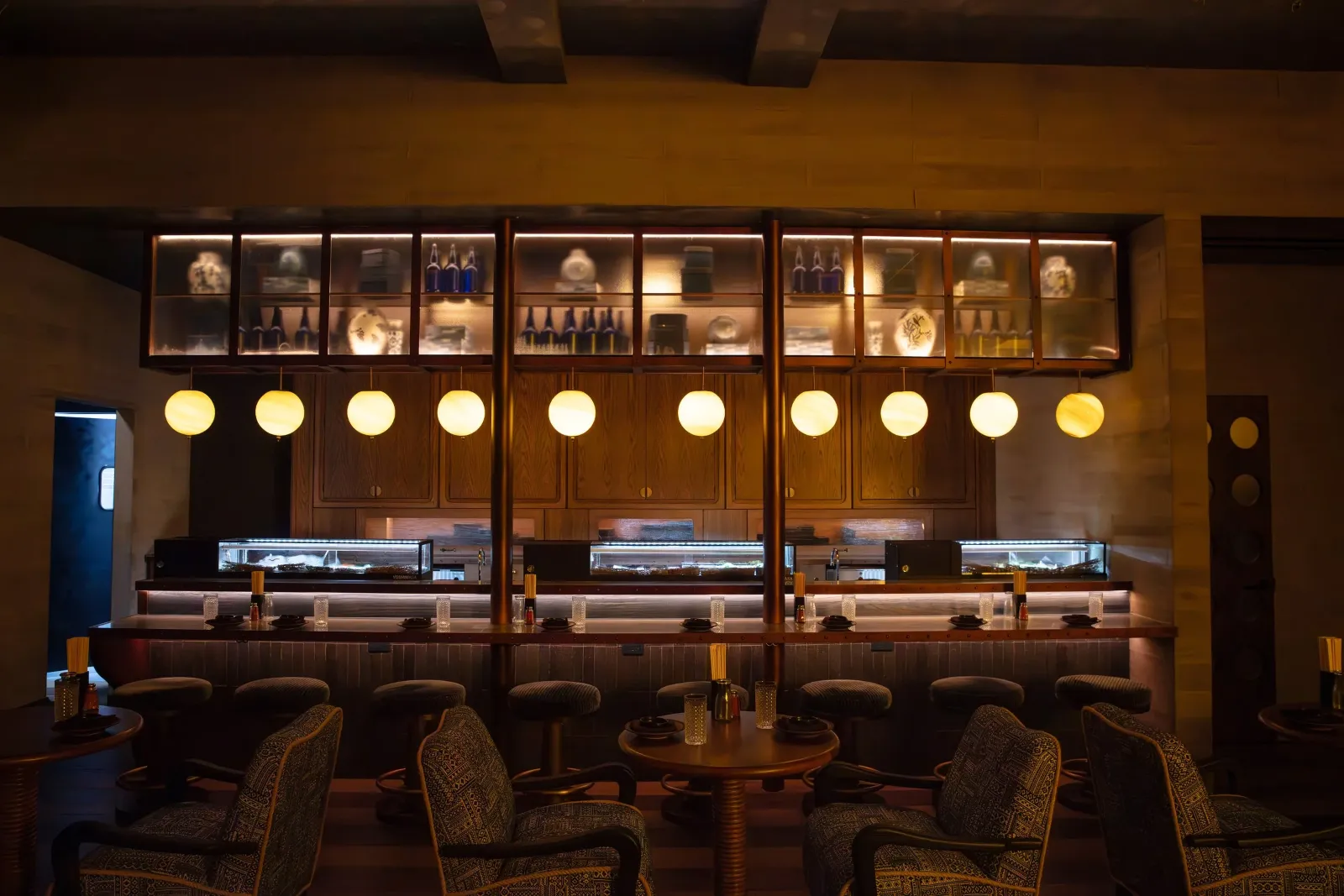 Interior of a restaurant with a bar area, illuminated pendant lights, and wooden furniture, featuring a display cabinet with decorative items on the wall behind the bar.
