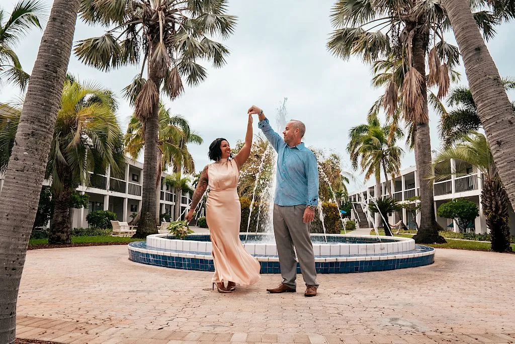 Two people are dancing by a fountain surrounded by palm trees.