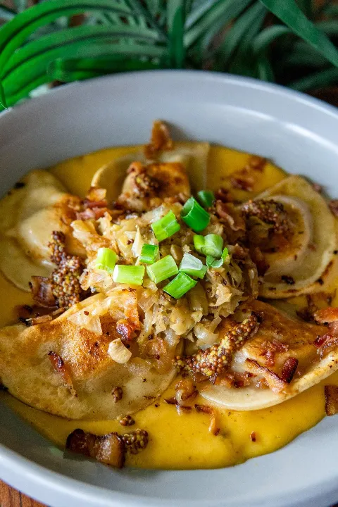 A bowl of pierogis topped with chopped green onions, grain mustard, bacon and beer cheese.