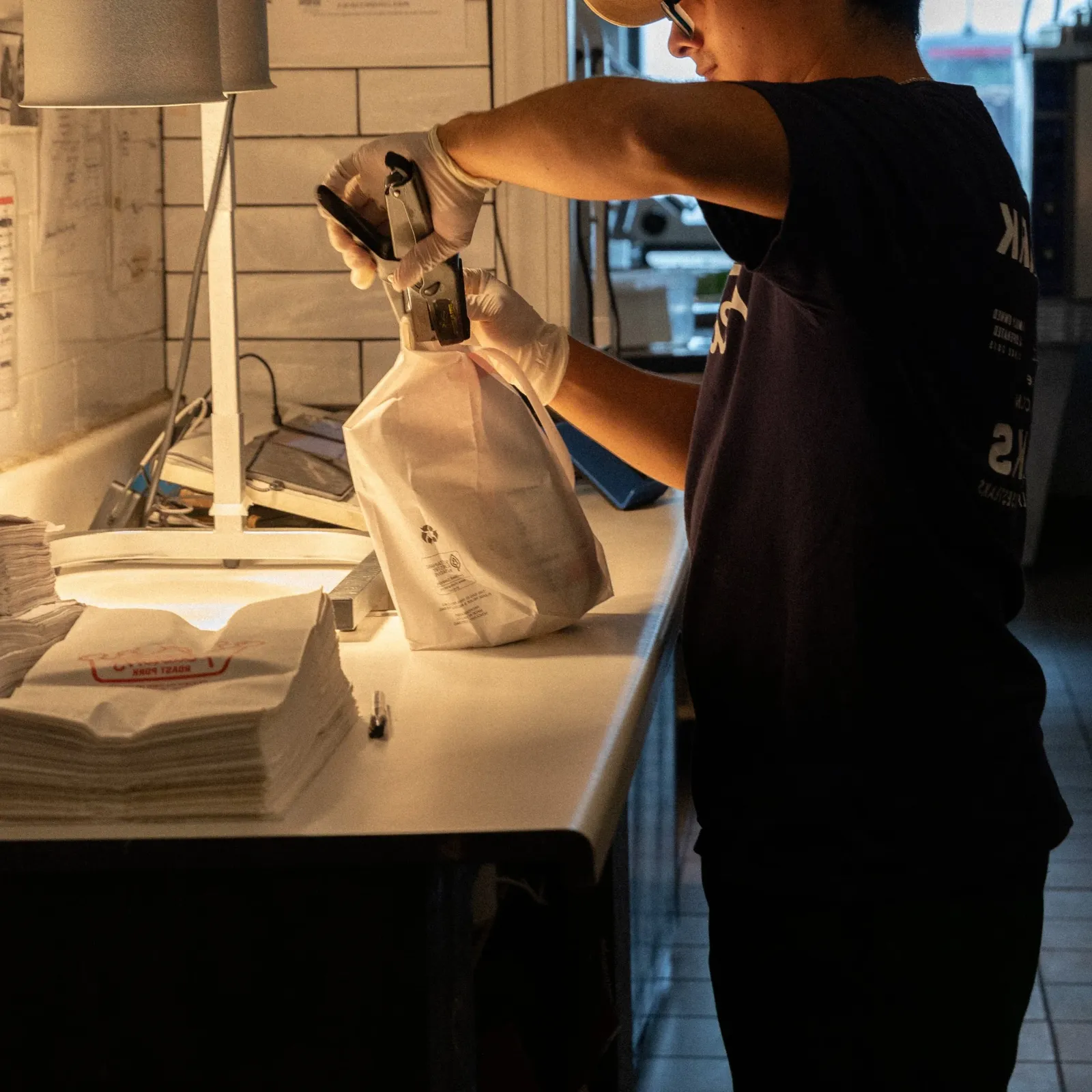 A person wearing gloves is sealing a paper bag with a heat sealer on a cluttered desk, with stacks of paper and a lamp nearby.