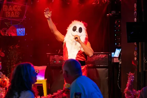 Person in a costume with a Jack Skellington mask, holding a microphone and dancing on a stage with red lighting at a bar or nightclub.