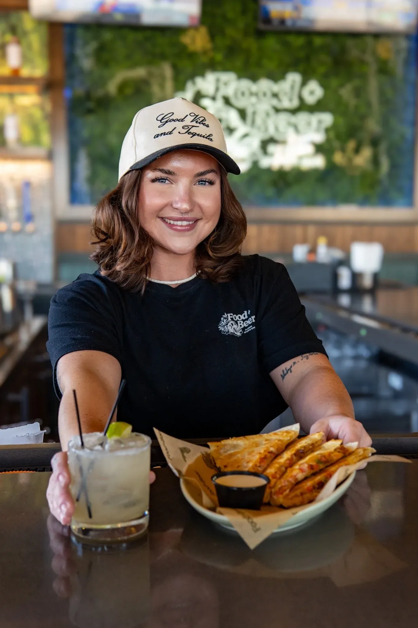A woman with shoulder-length brown hair wearing a beige baseball cap that says 'Good Vibes and Tequila', sitting at a restaurant table with a plate of sliced quesadillas, a drink with a lime wedge, an