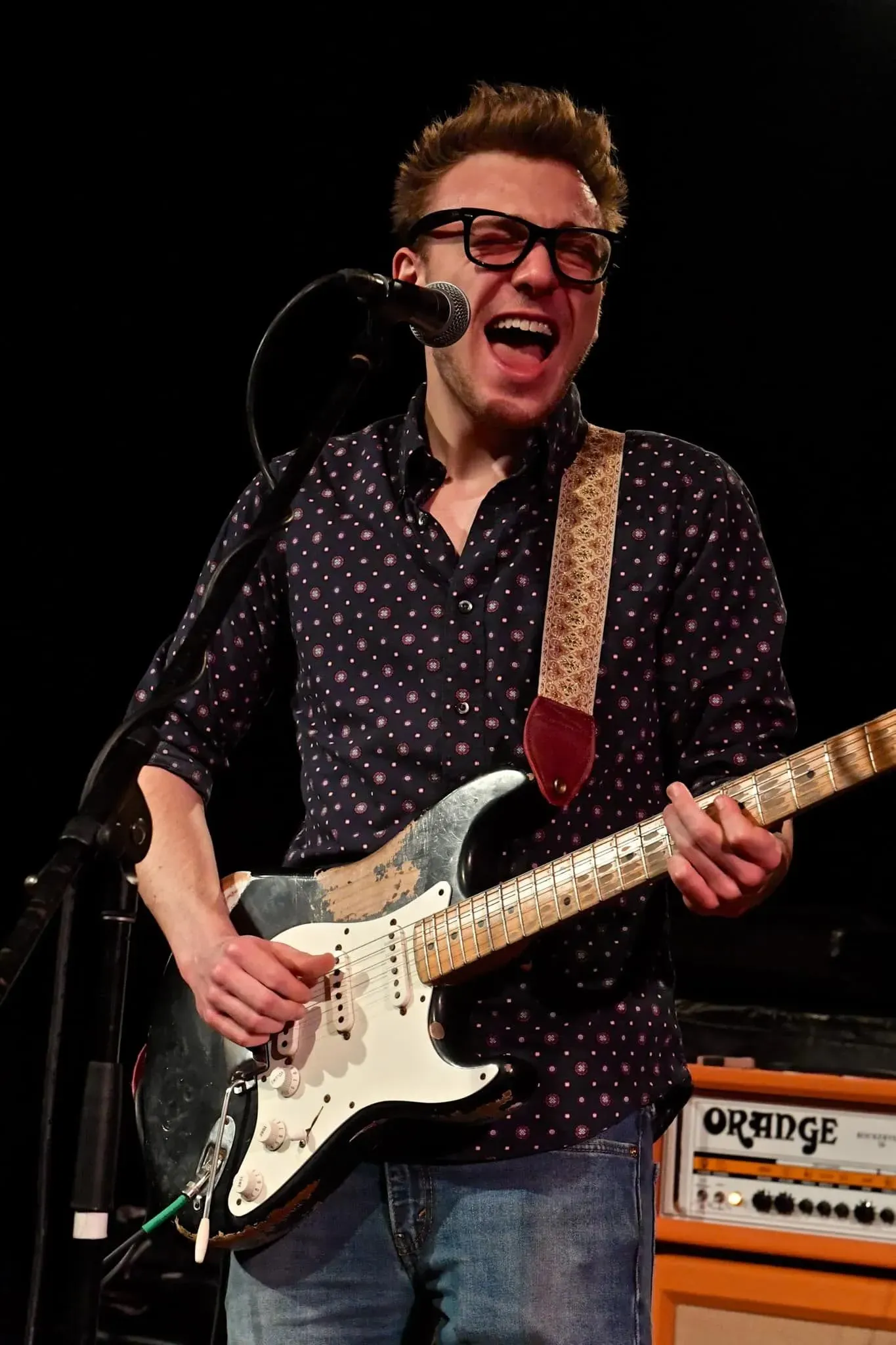 A musician wearing glasses and a patterned shirt sings while playing an electric guitar on stage near an Orange amplifier.