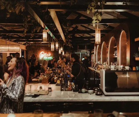 People socializing and bartending inside a dimly lit bar or nightclub with hanging pendant lights, a neon sign reading "Otto's HIGH DIVE," and a marble-topped bar counter filled with bottles and glass