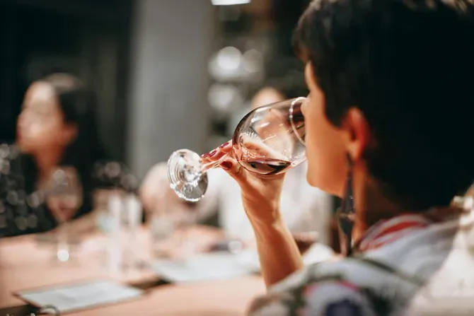 Woman sipping red wine at a table, blurry background with other people.