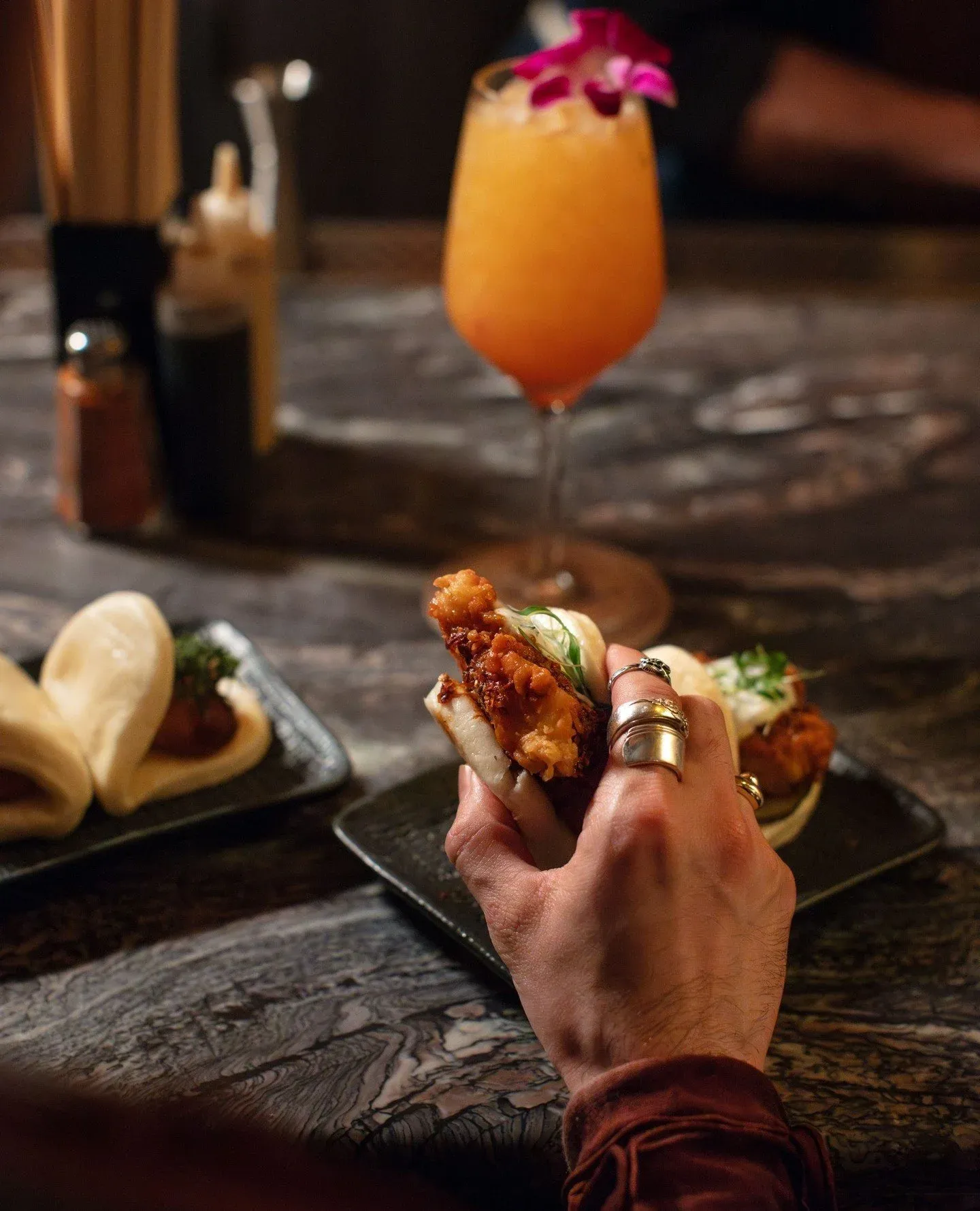 A hand holding a bao bun with fried chicken, with other bao on a black plate in the background. A pink flower cocktail in a tall glass is also visible on a wooden table.