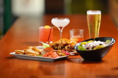 Assorted drinks and dishes on a wooden bar counter, including a salad in a black bowl, chicken wings, and various cocktails and beer.