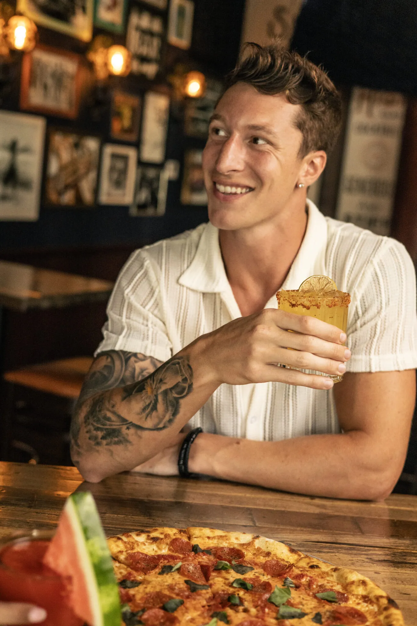 Tattooed man holding cocktail beside pizza at bar
