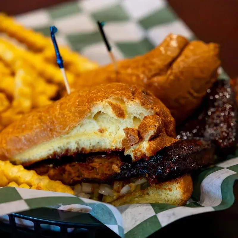 Close-up of a barbecue sandwich with a bun, pulled pork, and a side of french fries on checkered paper.