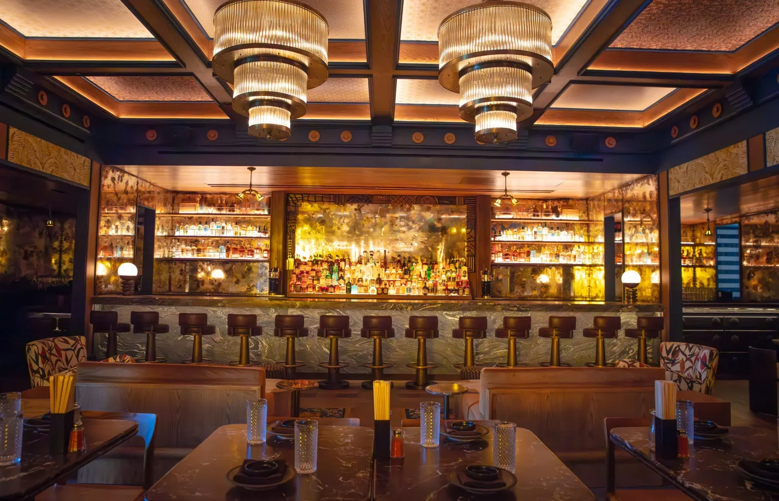Interior of a bar with a marble counter, multiple chairs, and a warmly lit ceiling. Behind the bar, various bottles of alcohol are displayed on illuminated shelves with decorative floral backgrounds. 