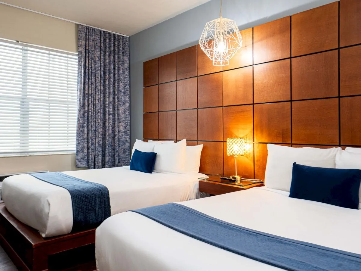 A hotel room with two queen beds, white linens, blue runners and pillows, a wooden paneled headboard, a nightstand lamp, and a window with curtains.