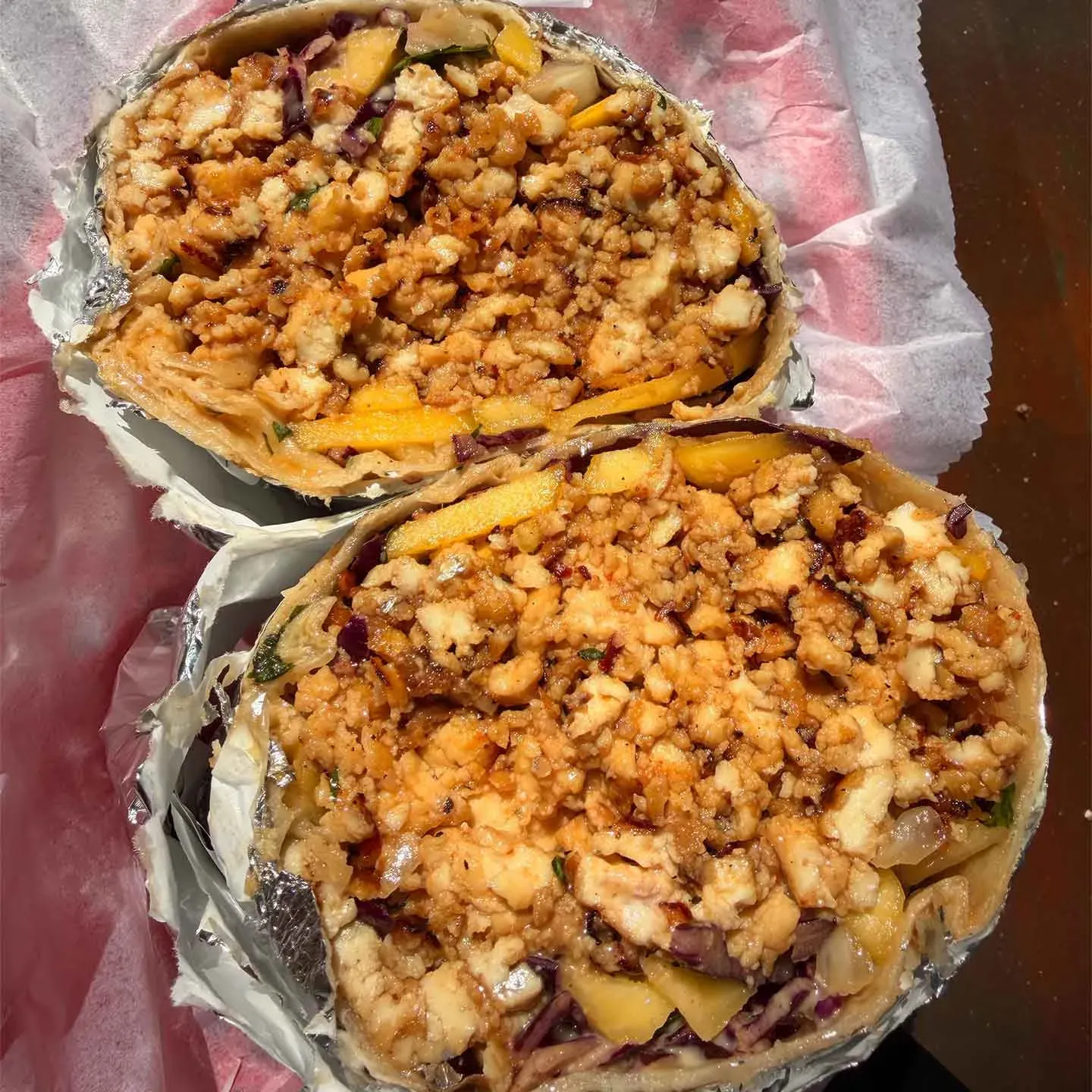Two halves of a wrap filled with crumbled tofu, vegetables, and mango, wrapped in foil on parchment paper.