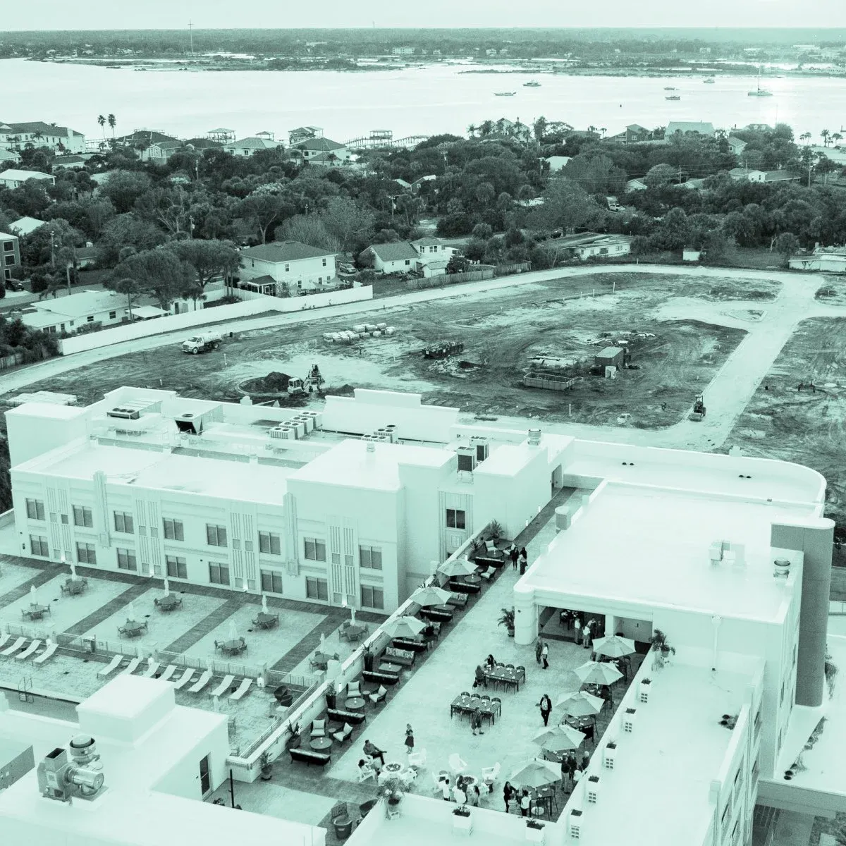 Aerial view of a large hotel or resort with a pool area, some outdoor seating, and an adjacent construction site with construction equipment and plots of land in development, near a waterfront with bo