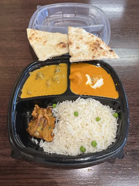 A meal consisting of basmati rice with peas, naan bread, vegetable curry, paneer curry, and a vegetable fritter in a black divided tray.