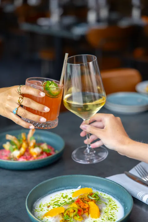 People toasting with a glass of white wine and a cocktail at a dining table, with plates of colorful food in the foreground.