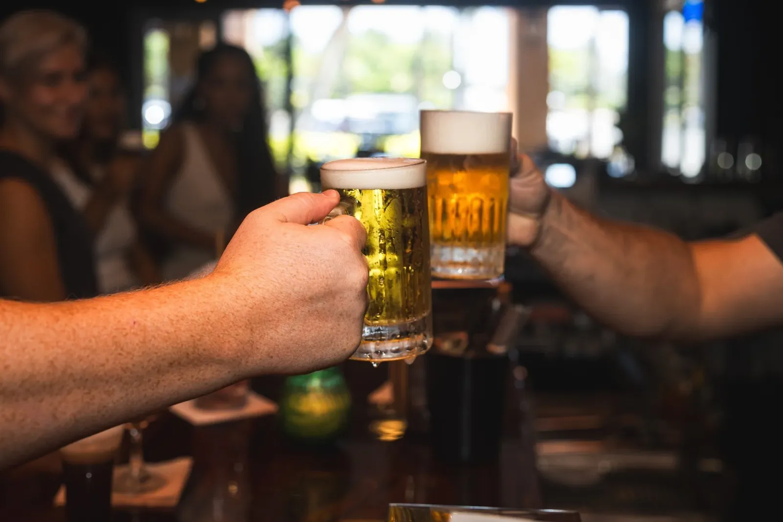 Two people clinking glasses of beer in a bar, with a group of women smiling in the background.